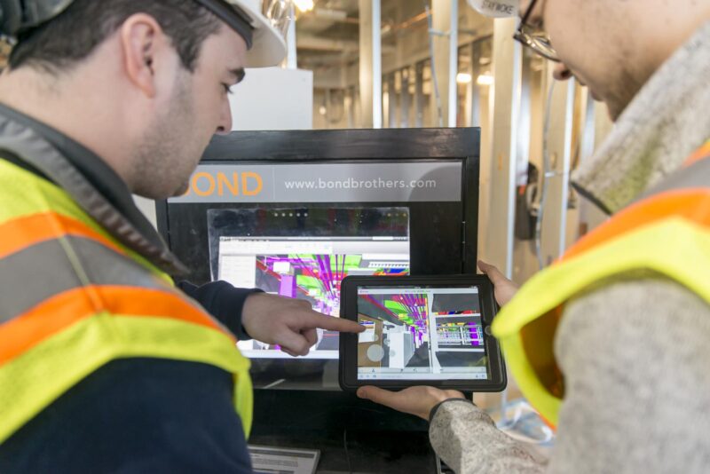 Two men in safety equipment in front of a BIM Box and holding and pointing at an iPad with Building Information Modeling/3D drawings on it.
