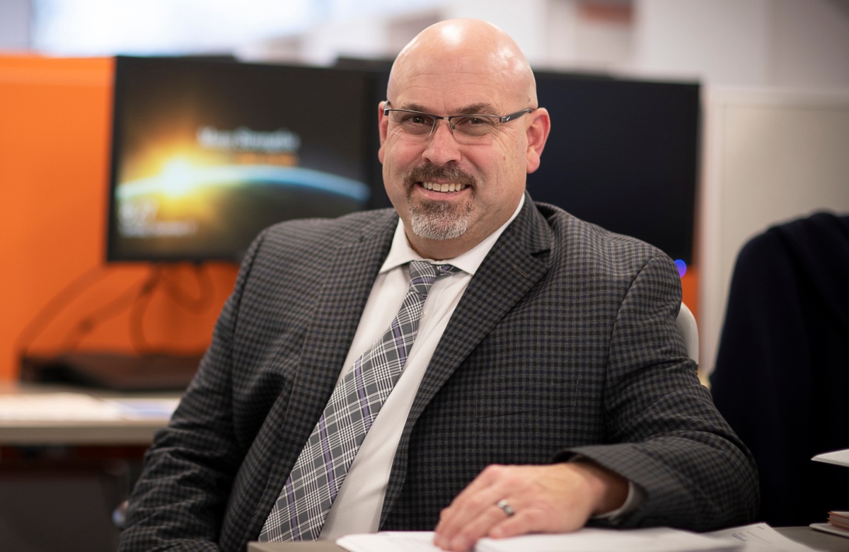 David Capaldo sitting at a desk with suit and tie on. Monitors are behind him.