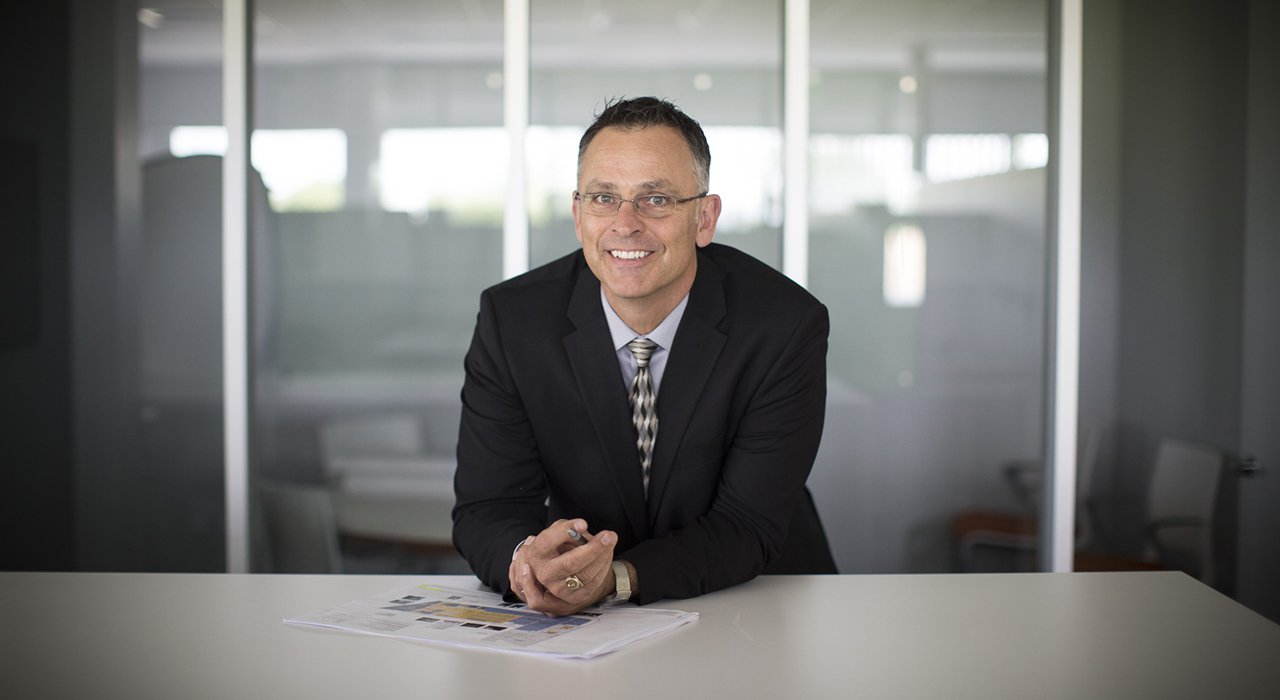 Man with suit and tie leaning on a counter in an office with conference room behind him