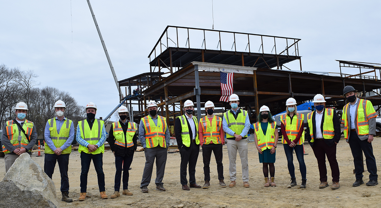 Town of Weymouth MA Maria Weston Chapman Middle School Topping Off - project team in hardhats, masks, and vests assembled in a line in front of a steel structure with the newly placed signed beam and an American flag