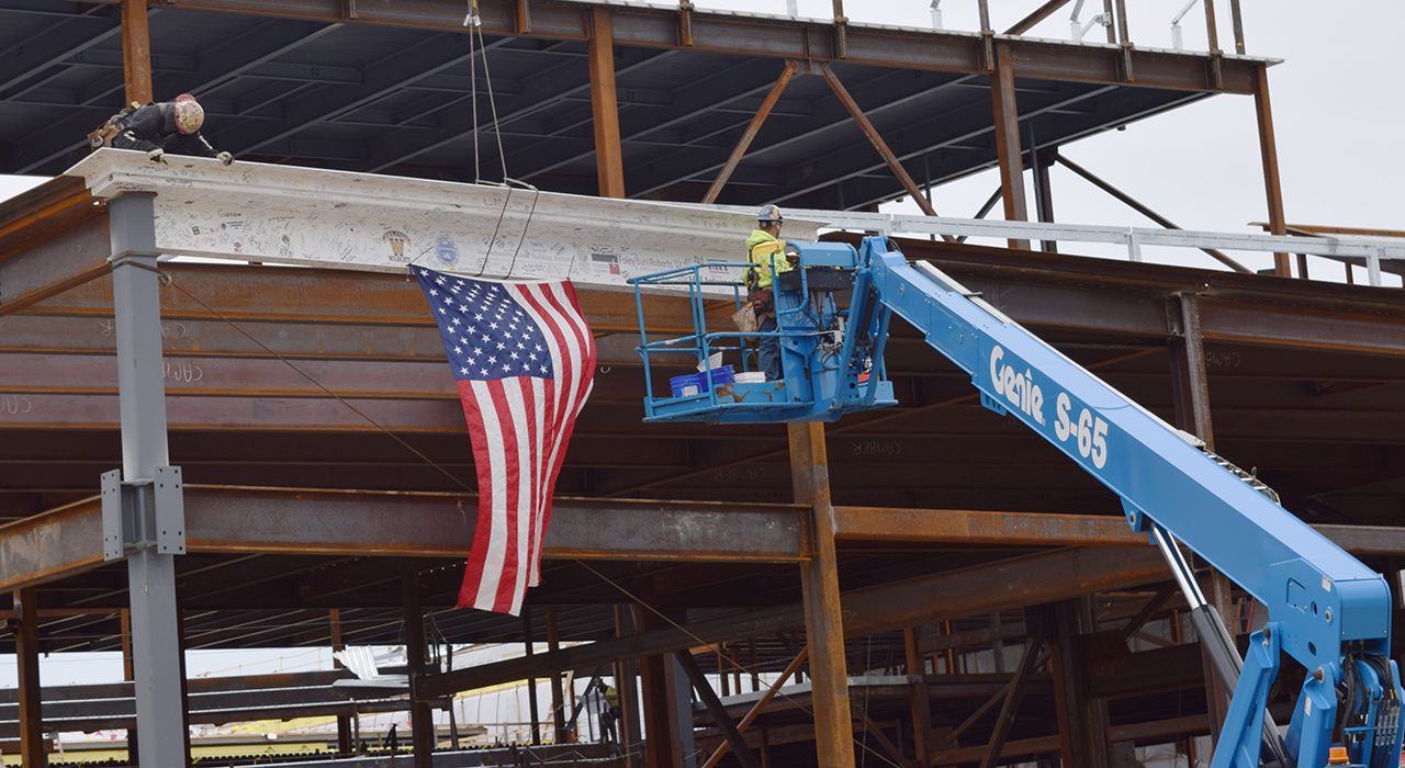 Town of Weymouth MA Maria Weston Chapman Middle School Topping Off - workers place topping off signed steel beam draped with an American flag. Blue crane in foreground.